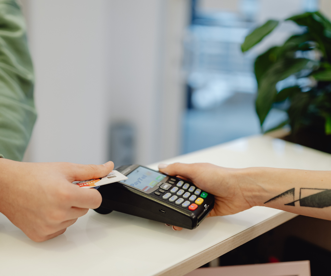 Divas rokas veic maksājumu, ievietojot kredītkarti maksājumu terminālī, ko tur otra roka. Uz termināļa ekrāna redzams uzraksts "PayTel".