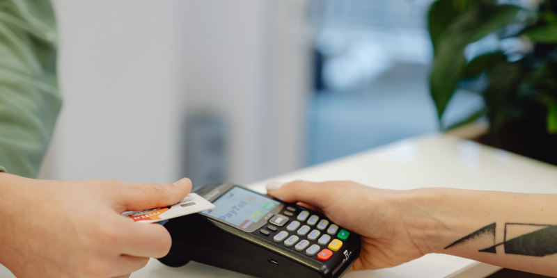 Divas rokas veic maksājumu, ievietojot kredītkarti maksājumu terminālī, ko tur otra roka. Uz termināļa ekrāna redzams uzraksts "PayTel".