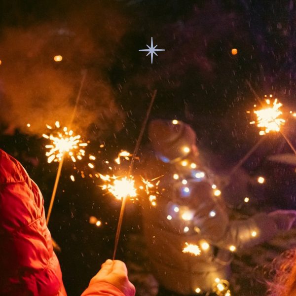 Tumšā naktī cilvēki tur rokās degošas brīnumsvecītes, kas rada zeltainas dzirksteles. Priekšplānā redzams cilvēks sarkanā jakā.