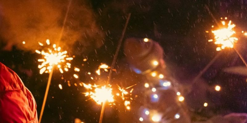 Tumšā naktī cilvēki tur rokās degošas brīnumsvecītes, kas rada zeltainas dzirksteles. Priekšplānā redzams cilvēks sarkanā jakā.