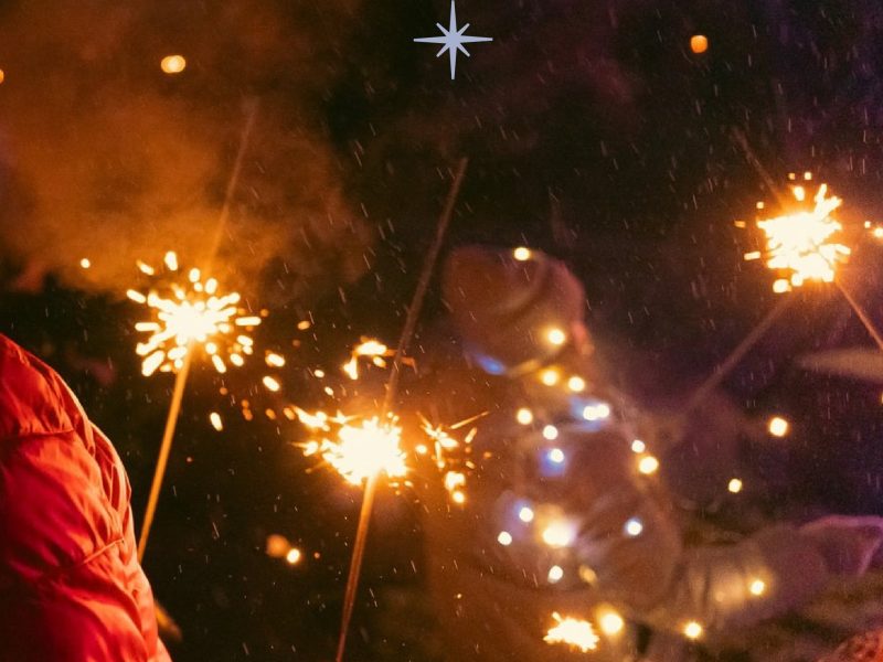 Tumšā naktī cilvēki tur rokās degošas brīnumsvecītes, kas rada zeltainas dzirksteles. Priekšplānā redzams cilvēks sarkanā jakā.
