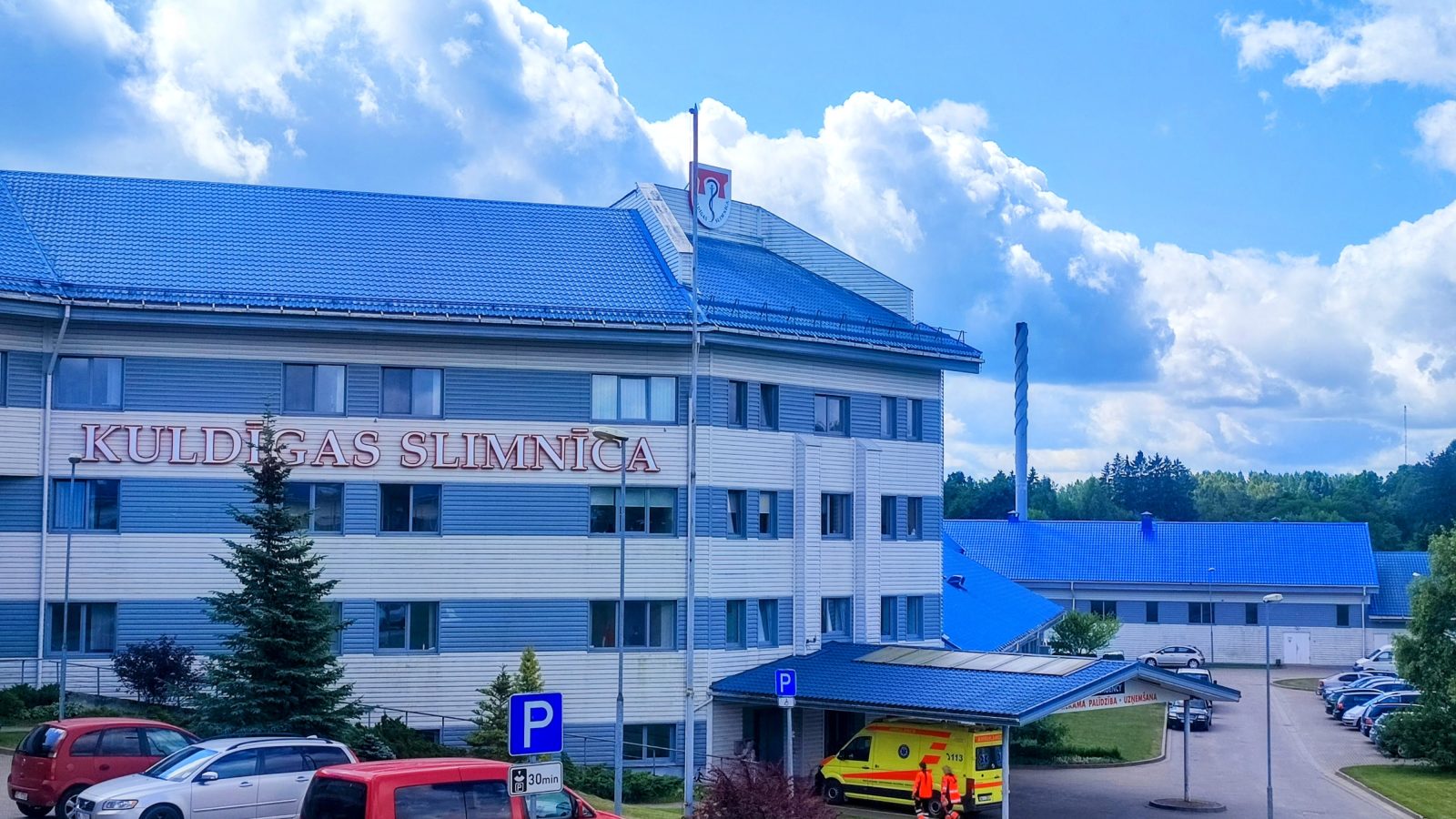 Kuldīgas slimnīcas galvenā ēka ar ziliem jumtiem un gaišām fasādēm. Uz ēkas ir uzraksts "KULDĪGAS SLIMNĪCA".