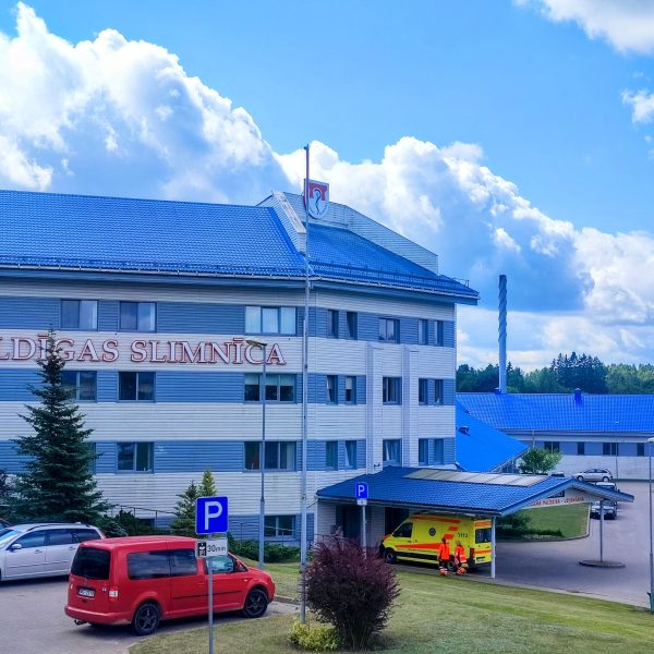 Kuldīgas slimnīcas galvenā ēka ar ziliem jumtiem un gaišām fasādēm. Uz ēkas ir uzraksts "KULDĪGAS SLIMNĪCA".