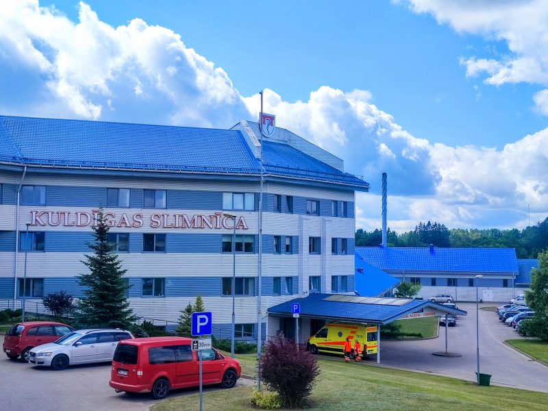 Kuldīgas slimnīcas galvenā ēka ar ziliem jumtiem un gaišām fasādēm. Uz ēkas ir uzraksts "KULDĪGAS SLIMNĪCA".