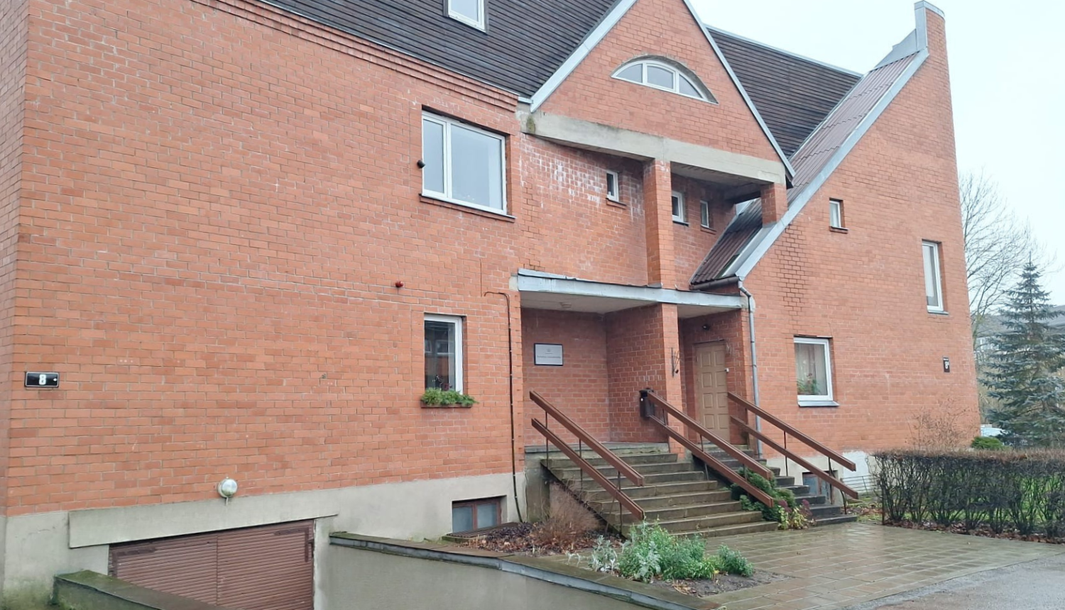 Sarkana ķieģeļu ēka ar stāviem jumtiem un dažādu formu logiem. Galvenā ieeja ar koka margām ved uz durvīm zem nelielas nojumes.