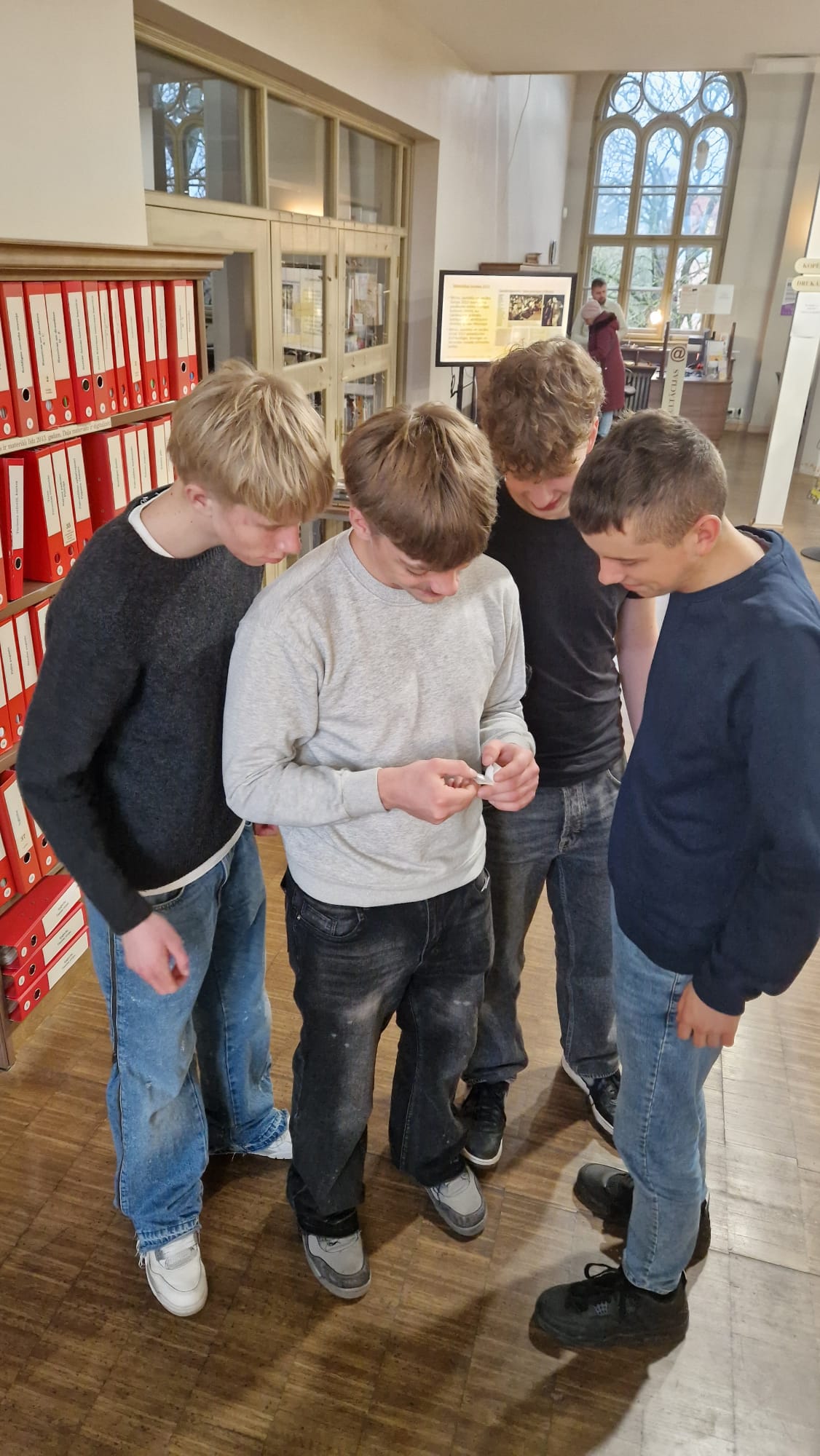 Četri jauniešitelpā pulcējas ap vienu, kurš pelēkā džemperī tur nelielu baltu priekšmetu, visiem uzmanīgi ieskatoties. Kreisajā pusē redzami plaukti ar sarkaniem sējumiem, bet fonā liels arkveida logs un displeja ekrāns.