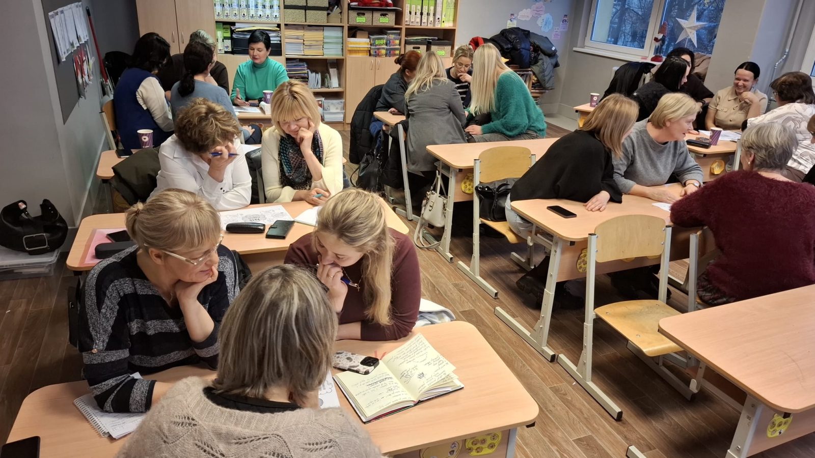 Sievietes sēž pie skolas galdiem klasē, iesaistoties diskusijās un pētot materiālus apmācību sesijā.