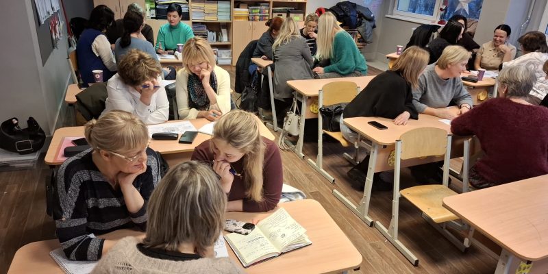 Sievietes sēž pie skolas galdiem klasē, iesaistoties diskusijās un pētot materiālus apmācību sesijā.