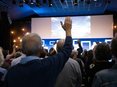 Cilvēku pūlis sanāksmju zālē, daudziem ir paceltas rokas. Viņi skatās uz lielu ekrānu. Mācības senioriem.