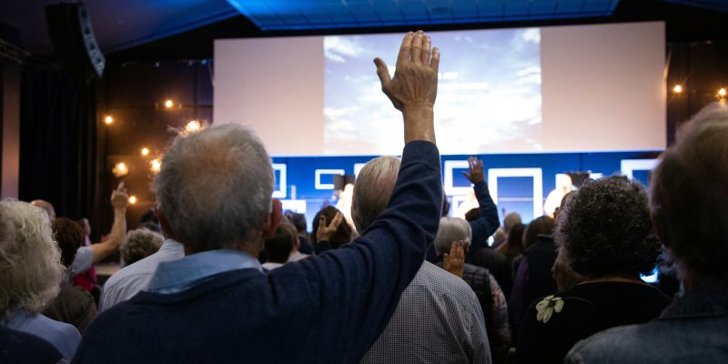 Cilvēku pūlis sanāksmju zālē, daudziem ir paceltas rokas. Viņi skatās uz lielu ekrānu. Mācības senioriem.