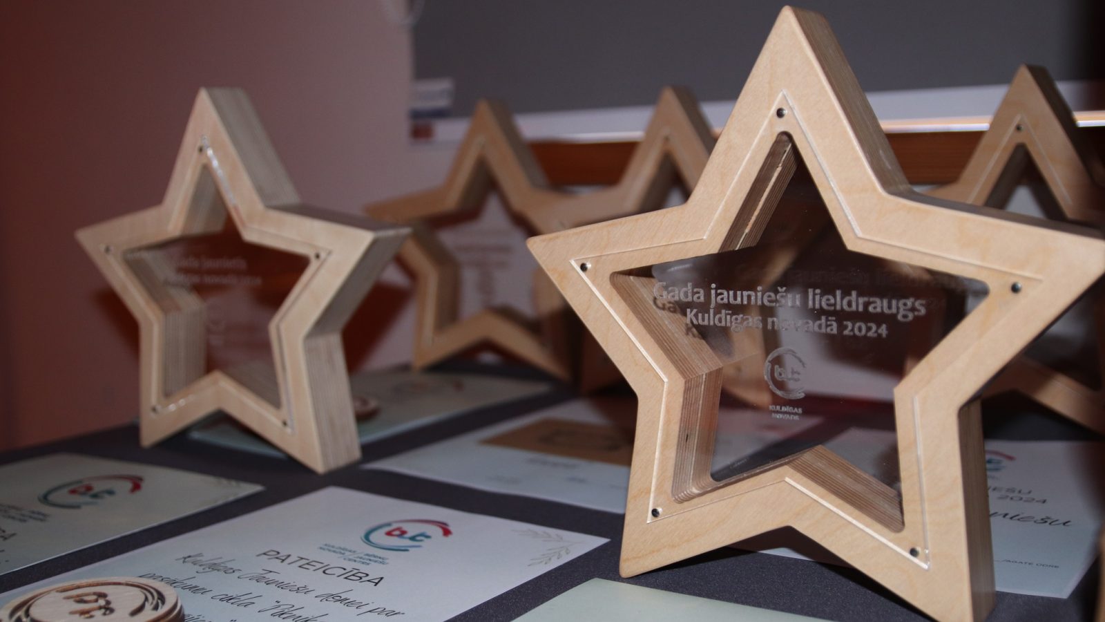 Vairāki koka zvaigznes formas apbalvojumi ar caurspīdīgiem centriem un sertifikāti uz galda. Uz priekšējā apbalvojuma ir uzraksts: "Gada jauniešu lieldraugs Kuldīgas novadā 2024".