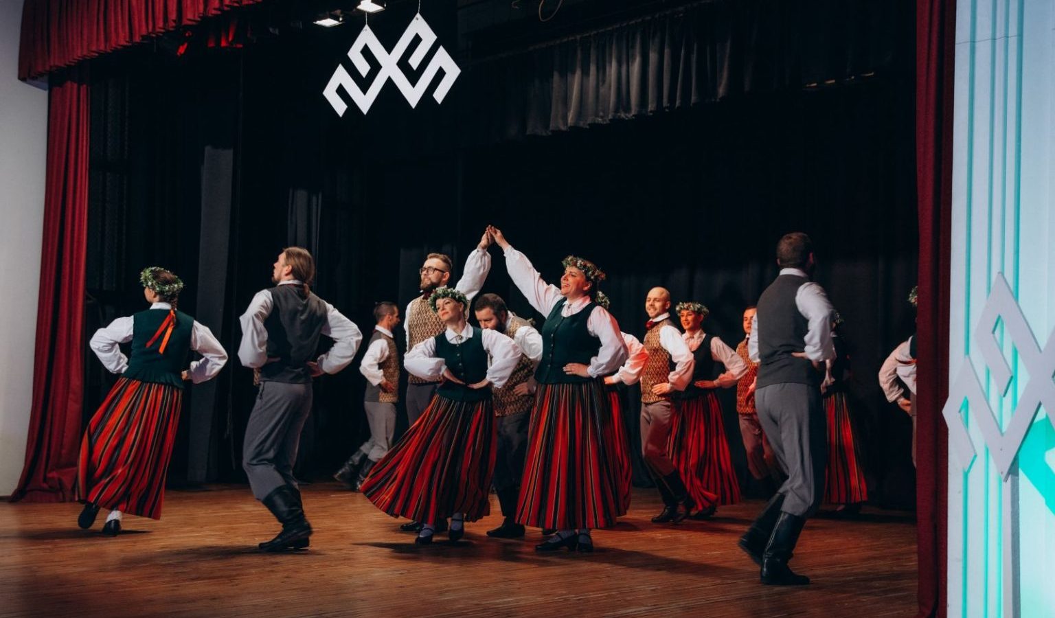 Folkloras dejotāji tradicionālos latviešu tērpos uzstājas uz skatuves. Sievietes valkā garus svītrainus svārkus, zaļas vestes un vainagus, vīrieši – pelēkas bikses un vestes. Aiz dejotājiem pie melnās sienas redzama liela balta Jumja zīme.