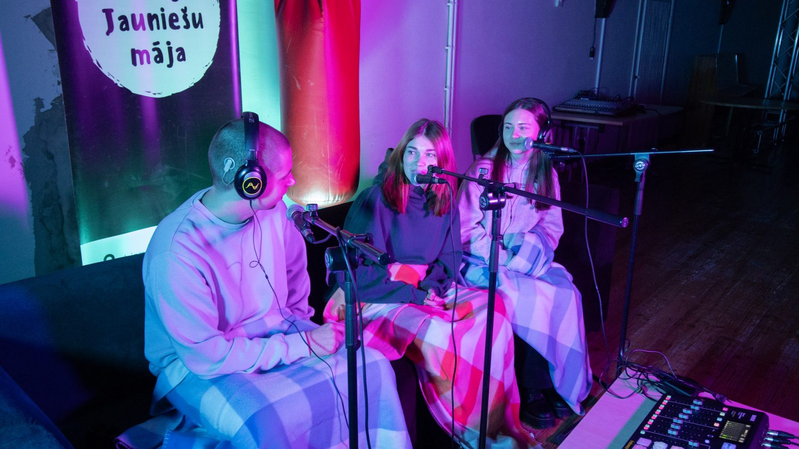 Trīs jaunieši, divas sievietes un vīrietis, sēž zem segām, runā mikrofonos un valkā austiņas, ierakstot podkāstu purpursarkanās un zilās gaismās. Fonā redzams plakāts ar uzrakstu "Kuldīgas Jauniešu māja".
