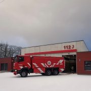 Sarkana ugunsdzēsēju mašīna ar baltām svītrām, uzrakstu "BLAŅES", emblēmu un numuru "402" novietota pie moderna ugunsdzēsēju depo ēkas, uz kuras fasādes redzami lieli sarkani cipari "112". Zeme ir apsnigusi.