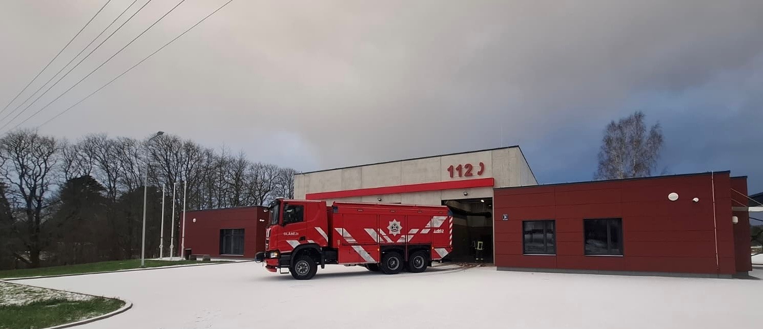 Sarkana ugunsdzēsēju mašīna ar baltām svītrām, uzrakstu "BLAŅES", emblēmu un numuru "402" novietota pie moderna ugunsdzēsēju depo ēkas, uz kuras fasādes redzami lieli sarkani cipari "112". Zeme ir apsnigusi.