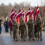 Vairāki vīrieši militārās maskēšanās formās soļo pa ielu, nesot paceltus Latvijas karogus. Fonā redzams orķestris