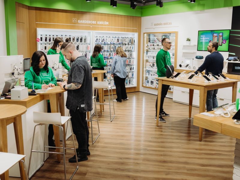 Moderns elektronikas veikals, kurā darbinieki zaļos džemperos apkalpo klientus.
