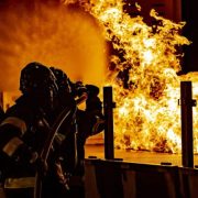 Divi ugunsdzēsēji aizsargtērpos dzēš lielu, spilgtu ugunsgrēku tumšā telpā, viens tur ugunsdzēsības šļūtenes uzgali.