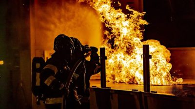 Divi ugunsdzēsēji aizsargtērpos dzēš lielu, spilgtu ugunsgrēku tumšā telpā, viens tur ugunsdzēsības šļūtenes uzgali.