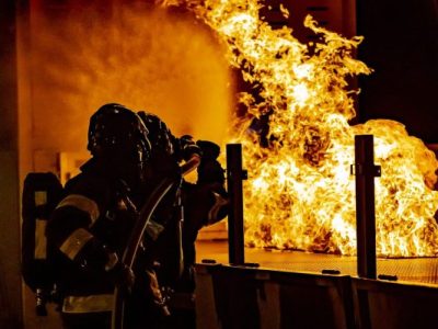 Divi ugunsdzēsēji aizsargtērpos dzēš lielu, spilgtu ugunsgrēku tumšā telpā, viens tur ugunsdzēsības šļūtenes uzgali.