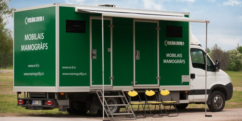 Zaļi-balts kravas auto ar numura zīmi HK-349, uz kura sāniem rakstīts "VESELĪBAS CENTRS 4" un "MOBILAIS MAMOGRĀFS", kā arī "www.vc4.lv" un "www.mamografija.lv", stāv ārā ar izvilktu nojumi, kāpnēm un trim dzelteniem saliekamiem krēsliem.