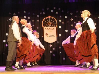 Pieaugušo tautas deju kolektīvs tradicionālos tērpos dejo uz skatuves. Dejotāji, vīrieši pelēkos uzvalkos un sievietes sarkanos rūtainos svārkos ar melnām vestēm, ir izvietoti divās rindās, sievietēm turot paceltus svārkus. Fonā tumšs aizkars ar izkārtotiem kompaktdiskiem un baneris, uz kura redzams uzraksts "KABILE VPDK Meždzirnas" un stilizēts koka rats.