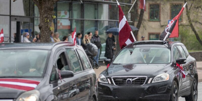 Mitrā ielā brauc divas melnas automašīnas, rotātas ar Latvijas karogiem. Priekšējais melnais apvidus auto uz jumta nes divus Latvijas karogus un sarkanmelnu karogu ar simbolu, uz tā sāniem ir uzraksts "Paldies visiem". Aizmugurējā automašīna ir daļēji redzama ar sarkanbaltsarkanu lentu. Fonā, uz ietves, stāv cilvēku pūlis, daži zem lietussargiem, vērojot notiekošo lietainā laikā.
