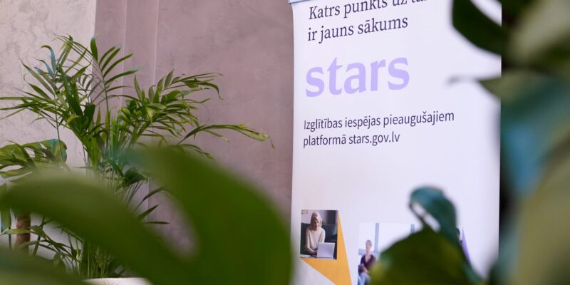 Balts stends ar tekstu "Katrs punkts uz taisnes ir jauns sākums", lieliem burtiem "stars" un "Izglītības iespējas pieaugušajiem platformā stars.gov.lv", redzams telpās starp zaļiem augiem.