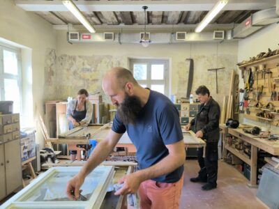 Trīs cilvēki – divi vīrieši un sieviete – restaurē logu rāmjus darbnīcā. Priekšplānā bārdains vīrietis tīra balta loga rāmi ar stiklu. Aizmugurē sieviete strādā pie koka rāmja, kamēr cits vīrietis apstrādā koka detaļas pie darbagalda. Telpai ir nolupušas sienas, atklātas griestu sijas un daudz darbarīku.
