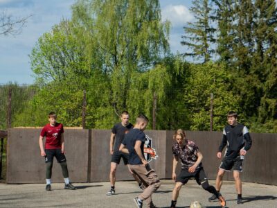 Pieci jauni vīrieši spēlē ielu futbolu uz āra asfalta laukuma ar koka sienām. Ēdolē notiks kausa izcīņa ielu futbolā