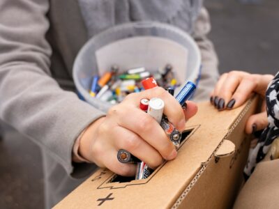 Tuvplānā redzamas personas rokas, kas nodod baterijas otrreizējai pārstrādei. Viena roka tur dažādas izlietotas baterijas, tostarp "Energizer" un "SWISS MADE" bateriju, un ievieto tās spraugā brūnā kartona kastē, kas paredzēta otrreizējai pārstrādei. Uz kastes ir pārsvītrotas atkritumu tvertnes simbols. Fonā spainis ir piepildīts ar citām izlietotām baterijām.