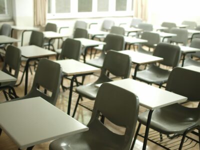 Tukša klase ar pelēkiem studentu krēsliem un gaišiem galdiem, kas izvietoti rindās. Telpu maigi apgaismo gaisma no logiem.