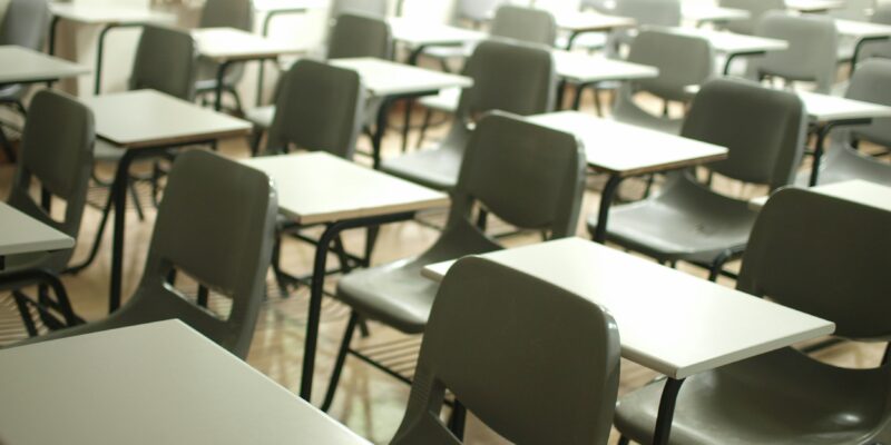 Tukša klase ar pelēkiem studentu krēsliem un gaišiem galdiem, kas izvietoti rindās. Telpu maigi apgaismo gaisma no logiem.