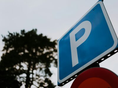 Zila taisnstūra stāvvietas zīme ar baltu burtu "P", redzama no apakšas pret pelēkām debesīm, ar izplūdušu tumšu koku fonā un daļēji redzamu sarkanu zīmi zem tās.