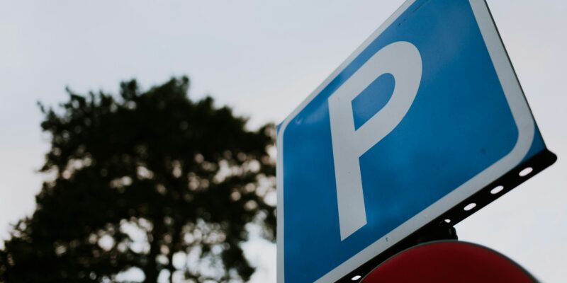 Zila taisnstūra stāvvietas zīme ar baltu burtu "P", redzama no apakšas pret pelēkām debesīm, ar izplūdušu tumšu koku fonā un daļēji redzamu sarkanu zīmi zem tās.