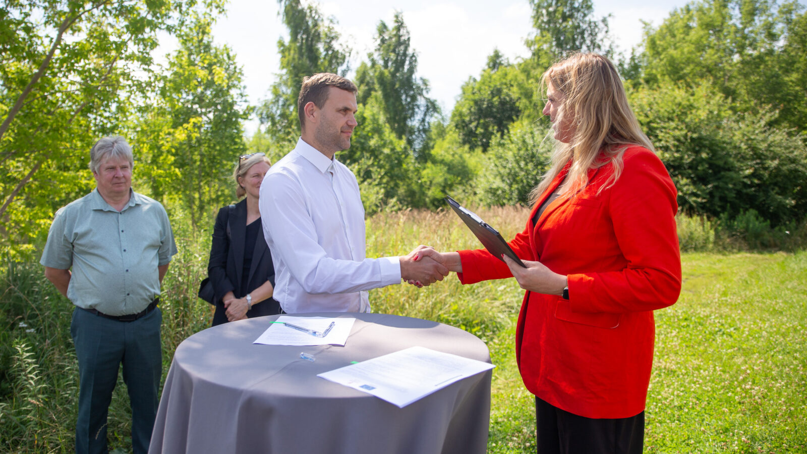 Vīrietis baltā kreklā un sieviete sarkanā žaketē - Kuldīgas novada domes priekšsēdētāja - paspiež rokas brīvā dabā, stāvot pie apaļa galda ar dokumentiem, tostarp vienu ar Eiropas Savienības simbolu.