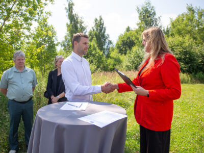 Vīrietis baltā kreklā un sieviete sarkanā žaketē - Kuldīgas novada domes priekšsēdētāja - paspiež rokas brīvā dabā, stāvot pie apaļa galda ar dokumentiem, tostarp vienu ar Eiropas Savienības simbolu.