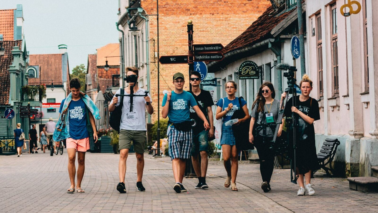 Seši jauni cilvēki iet pa bruģētu pilsētas ielu. Daži valkā T-kreklus ar uzrakstu "MEDIA SHARKS", un viens valkā T-kreklu ar uzrakstu "BAUNPILS". Divi no viņiem nes mediju aprīkojumu: viens tur kameras stabilizatoru, otrs – lielu statīvu ar mikrofonu. Fona ēkām ir vecas fasādes, un uz vienas ir uzraksts "SALONFRIZĒTAVA RITA anno 1964". Ielu zīme norāda uz "Annas baznīca 0.3", "WC 0.1", "Viegli uzliekas mašīna" un "BAUNPILS".