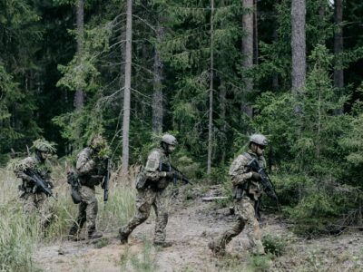 Četri karavīri maskēšanās tērpos ar šautenēm un Latvijas karoga ielāpiem uz pleciem pārvietojas pa mežu.
