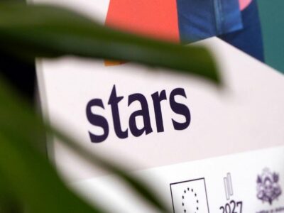 Balts plakāts vai stends ar vārdu "stars" tumši violetā krāsā. Zem tā redzams ES karoga logo, skaitlis "2027" un divi ģerbonim līdzīgi logo. Daļēji aizsegts ar zaļu augu lapām.